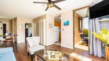 A modern living room with a glass coffee table, a ceiling fan, and a view of the kitchen and outdoor patio.