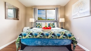 A bedroom with a bed, a ceiling fan, a window with curtains, a nightstand, a lamp, and a painting on the wall. - Kahana Manor 418 1 Bedroom Condo by RedAwning (Lahaina)