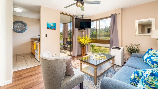 A living room with a sofa, armchair, coffee table, TV, and a ceiling fan. - Kahana Manor 418 1 Bedroom Condo by RedAwning (Lahaina)