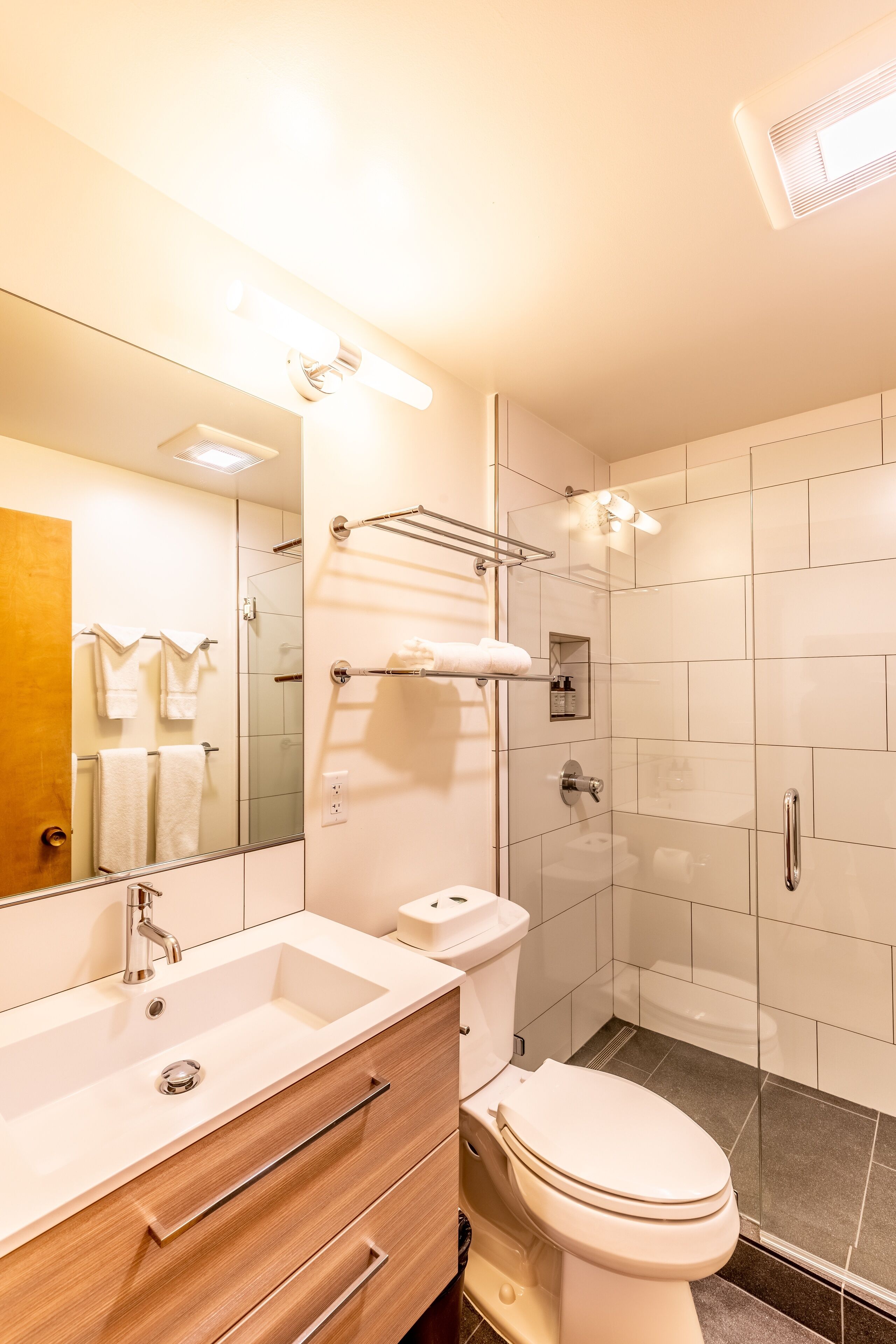 A modern bathroom with a white sink, vanity, and toilet. There is a glass shower enclosure and a mirror above the sink.