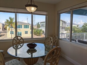 A dining area with a glass table and chairs, a hanging light fixture, and large windows offering views of neighboring buildings and palm trees.