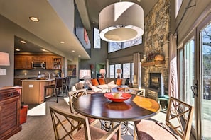 A spacious living area with a round wooden table, comfortable seating, a stone fireplace, and large windows offering a view of the outdoors.