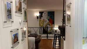 Um corredor com quadros nas paredes, um sofá de couro marrom, uma sala de jantar com mesa e cadeiras, e uma grande pintura abstrata na parede.