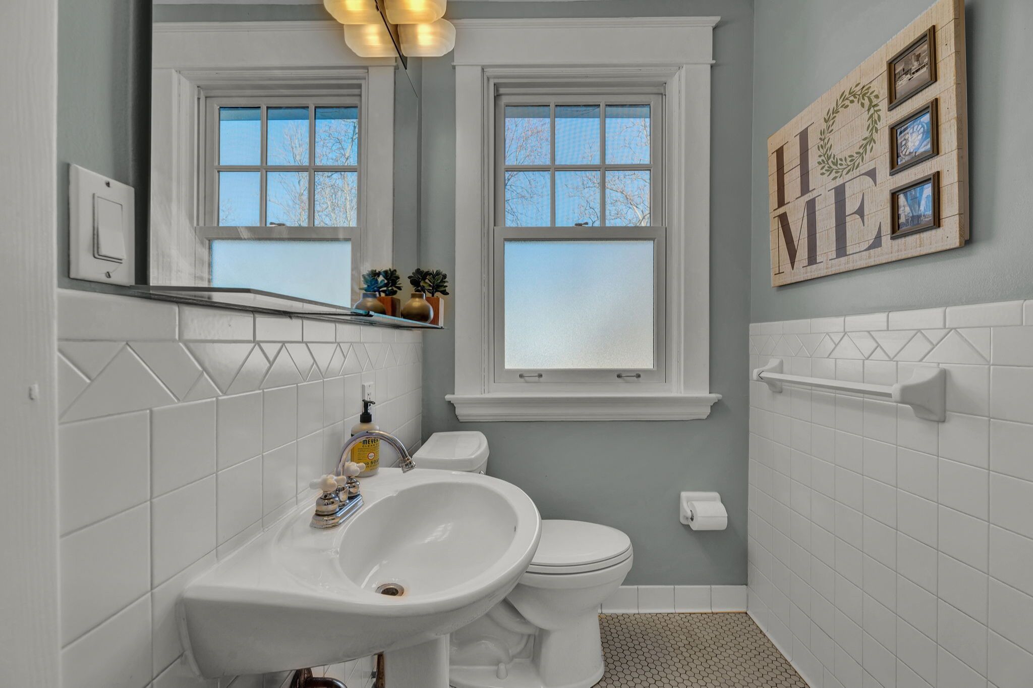 Un baño con un lavabo, inodoro y tocador blancos, dos ventanas y un letrero con la palabra 'HOME' en la pared.
