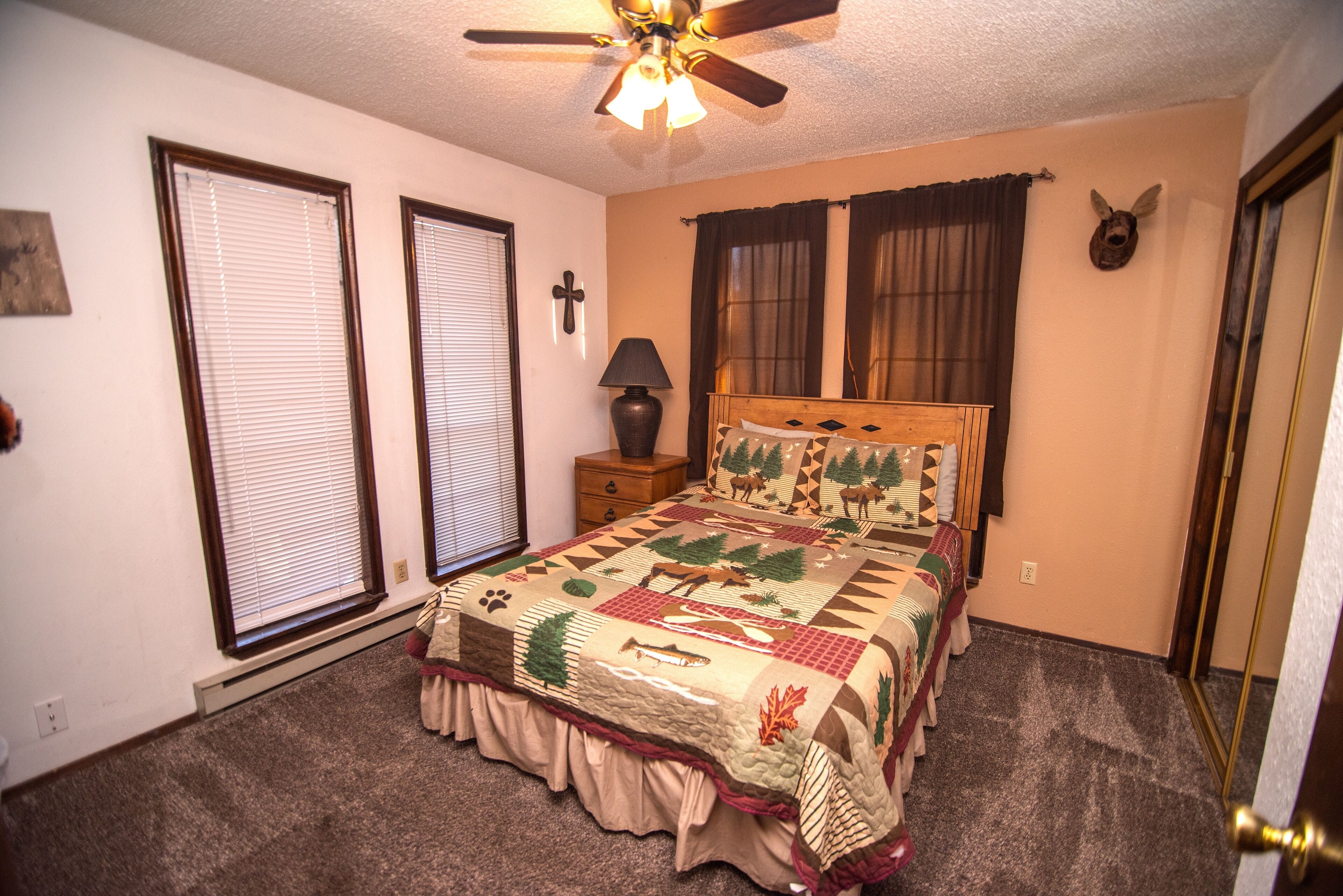 A bedroom with a bed, a ceiling fan, a nightstand, a lamp, and a wall-mounted deer head decoration.