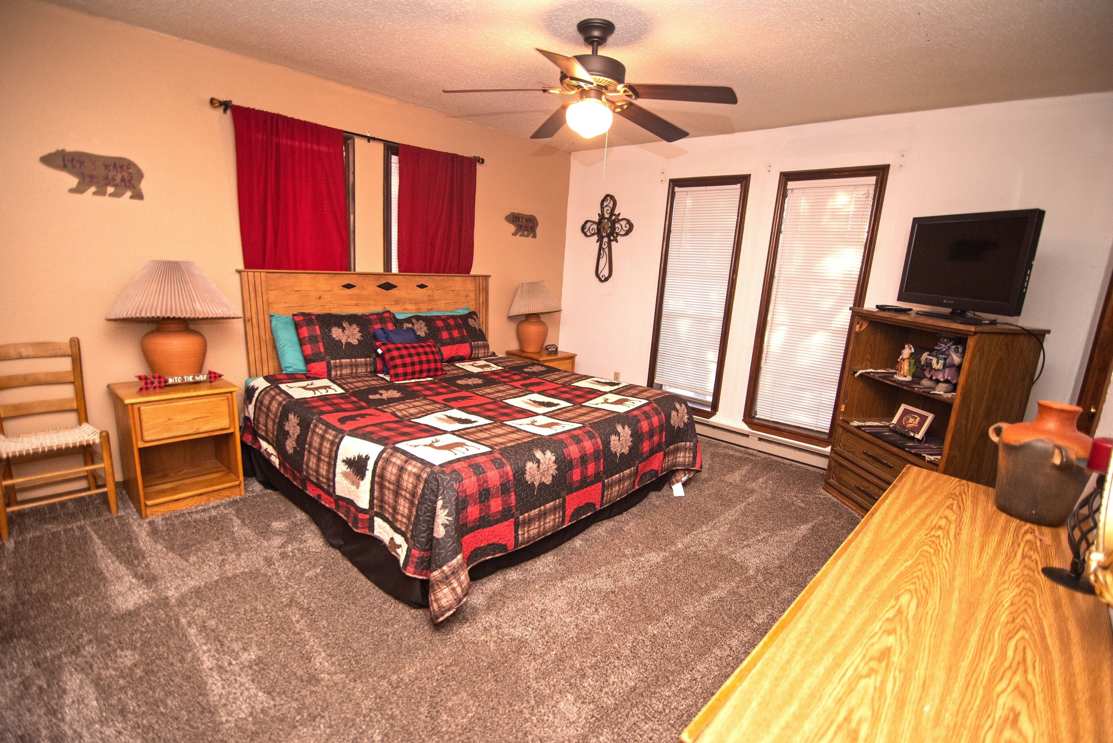 A bedroom with a bed, a ceiling fan, a TV, a wooden nightstand, a lamp, a chair, and a dresser.