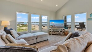 A spacious living room with a large window overlooking a beach, a comfortable sofa, and a television on a wooden stand. - The Shore House 3 Bedroom Home by RedAwning (Port Bolivar)