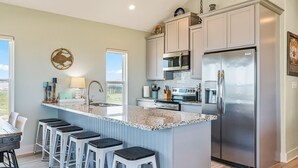 A modern kitchen with a central island, stainless steel appliances, and a view to the outdoors. - The Shore House 3 Bedroom Home by RedAwning (Port Bolivar)