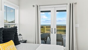 Un dormitorio con una cama, una mesita de noche, una lámpara y vistas a la playa a través de una puerta corrediza.