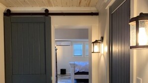 A hallway with a wooden ceiling, a blue door, and a patterned floor leading to a bedroom with a bed and a nightstand.