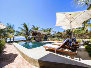 A poolside area with lounge chairs and umbrellas, surrounded by palm trees and a view of the ocean.