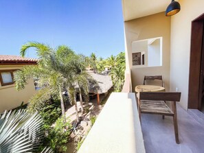 A balcony with a view of a tropical garden, a wooden chair, and a thatched-roof structure.