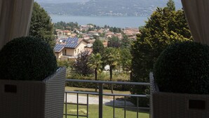 A view from a balcony with a railing, overlooking a lake, mountains, and a town with buildings and greenery. - Wonderful private villa with hot tub, sauna, private pool, WIFI, A/C, TV, patio and panoramic view (Baveno)