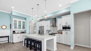 Cocina moderna con armarios blancos, una isla central con taburetes de bar, electrodomésticos de acero inoxidable e iluminación colgante.