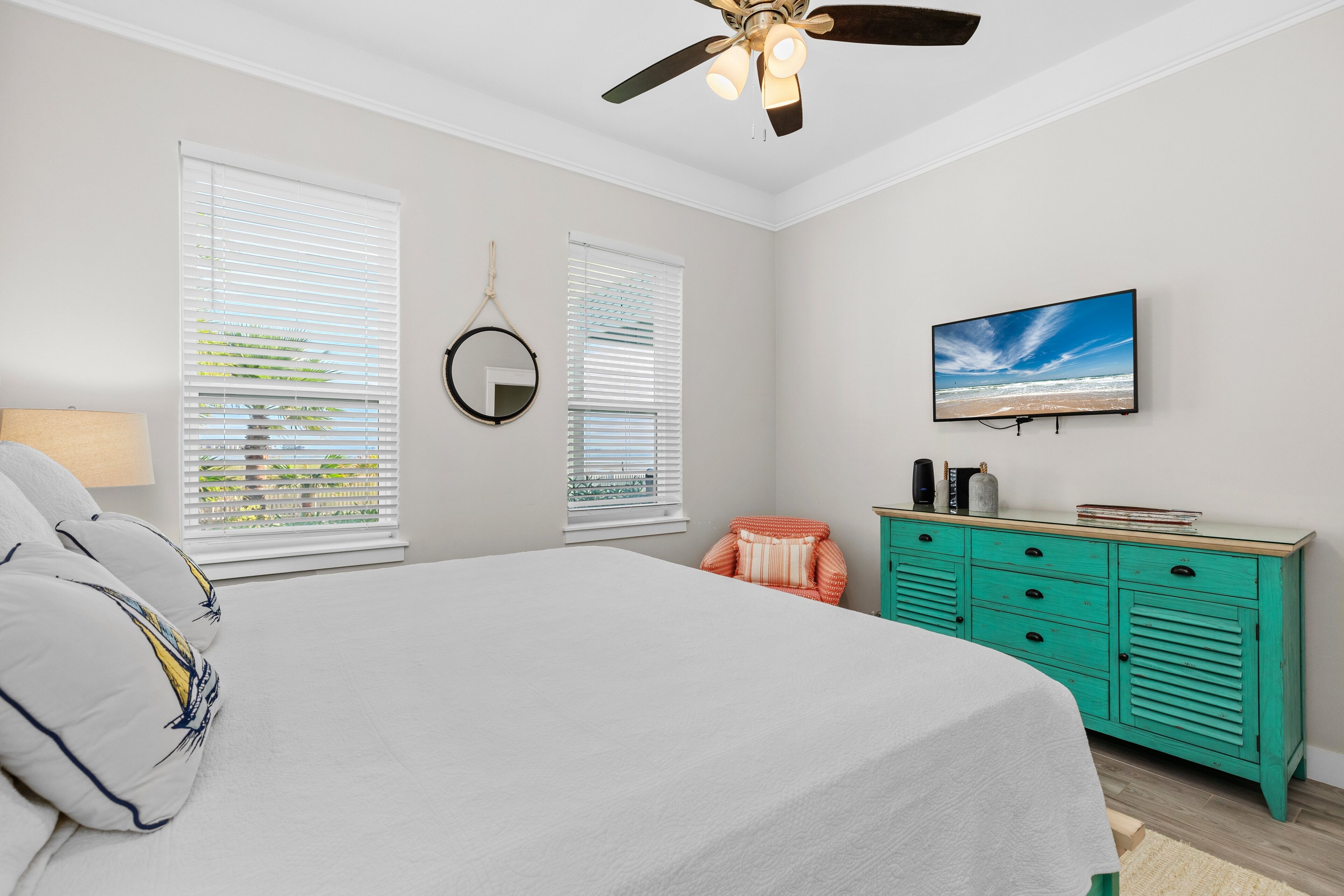 A bedroom with a bed, two windows with blinds, a ceiling fan, a television, a dresser, and a mirror.