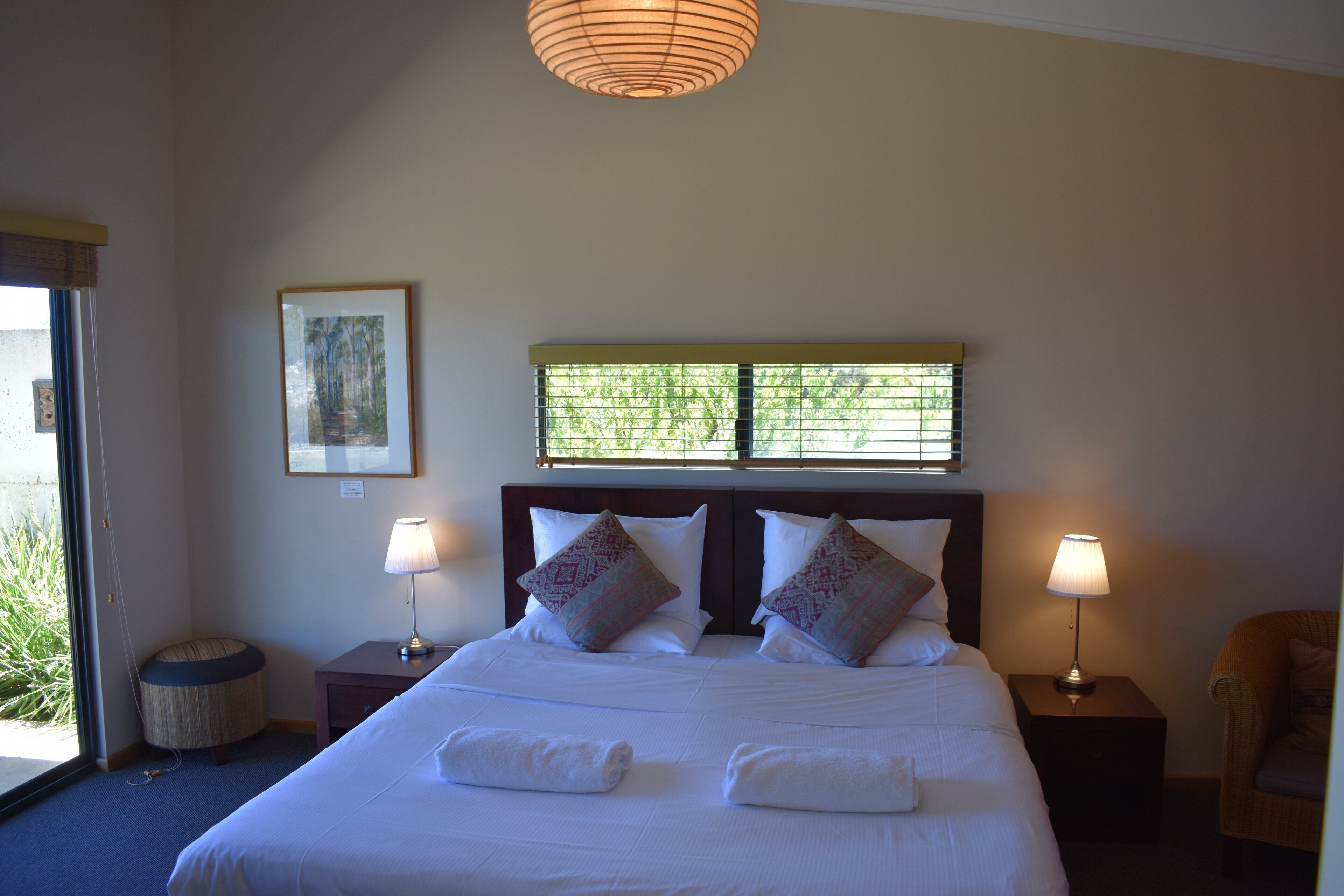 A hotel room with a large bed, two bedside lamps, a small round ottoman, a framed picture on the wall, and a window with blinds.