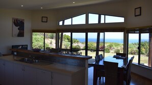 Una cocina moderna con una isla grande, zona de comedor con sillas y ventanas amplias con vista al océano.