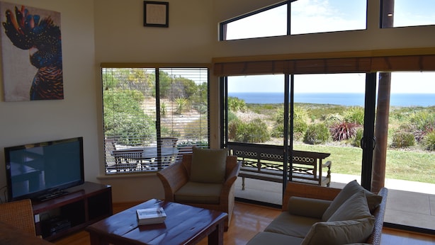 Sala de estar con televisor, sofá, mesa de centro y una silla. Hay un ventanal con vistas al océano y follaje.