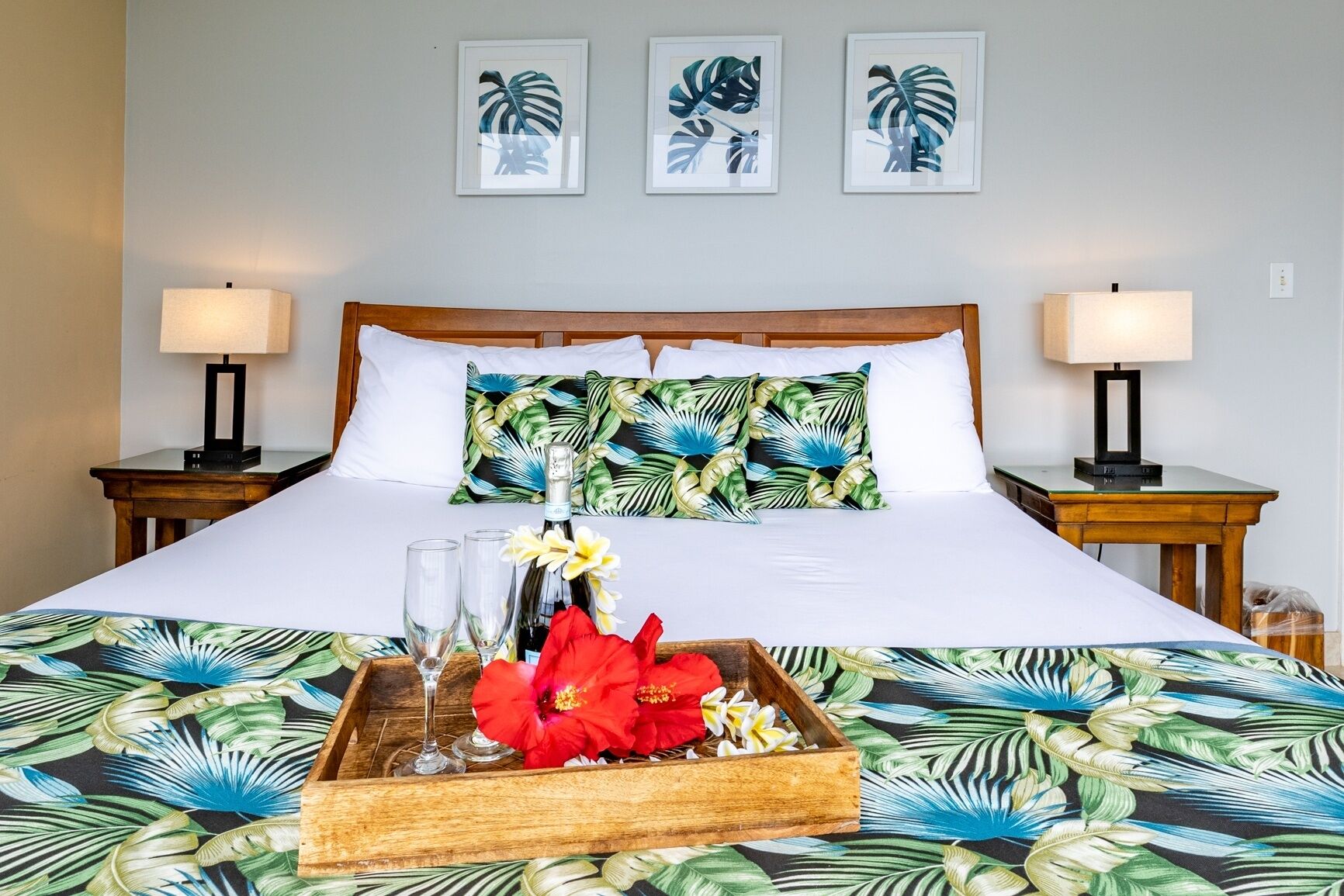 A hotel room with a bed, two bedside tables, and three framed tropical leaf artworks on the wall.