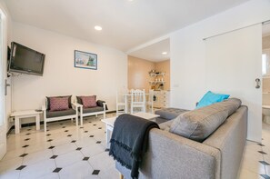Una sala de estar con un sofá, una mesa de centro, un televisor montado en la pared y una cocina visible a través de una puerta abierta.