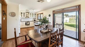 A dining area with a wooden table and chairs, a kitchen in the background, and a view through a glass door. - Ithaca Cottage by Your Innkeeper Mudgee (Frog Rock)