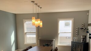A dining area with a wooden table and chairs, pendant lights, a radiator, and a cabinet with kitchen items. - Cottage Just 2 Blocks to Beach and Town (East Tawas)