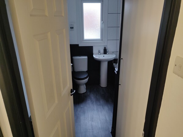 A narrow hallway leading to a bathroom with a toilet, sink, and window. - St Neots Riverside Park  1-Bed Top-Floor Apartment (Saint Neots)