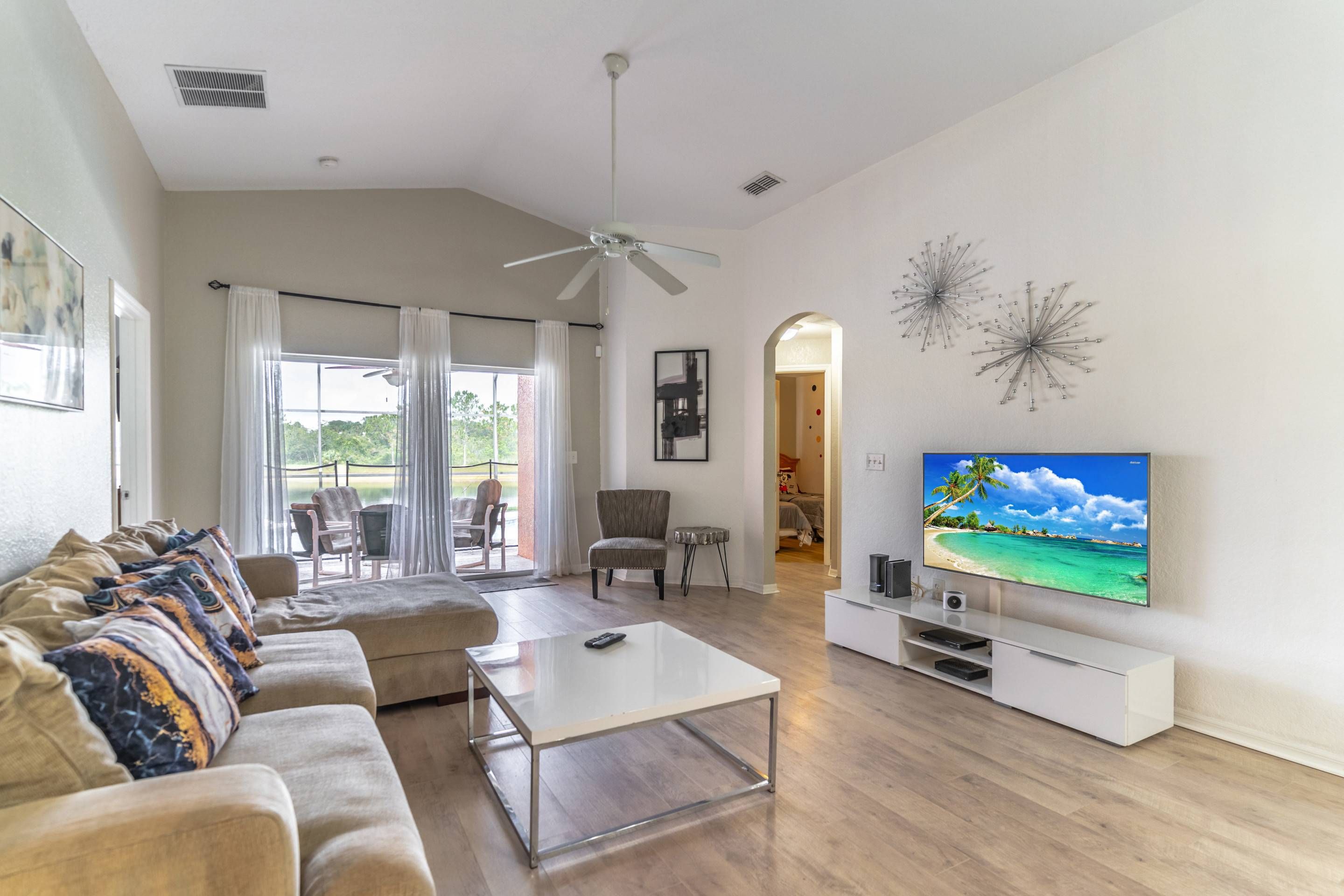 A spacious living room with a large flat-screen TV, a ceiling fan, and a view of the outdoors through large windows.