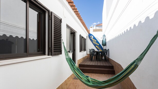Un estrecho pasillo exterior con una hamaca, un patio con sillas y un letrero azul con texto.