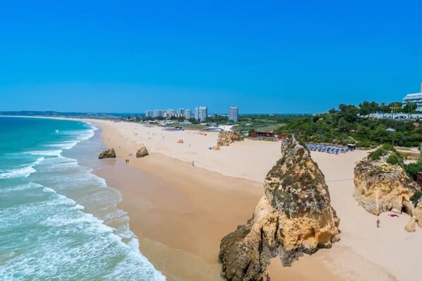 A beach with rocky formations, a clear blue sky, and buildings in the background.