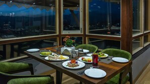 A dining area with a table set for two, a vase of flowers, and a view of the city at night. - Sumitel Vista 800m from Mall Road (Darjeeling)
