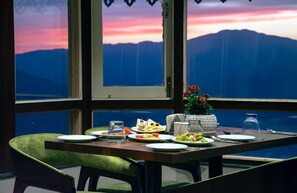 Une table à manger dressée avec des assiettes, des couverts et un vase de fleurs, avec une vue sur les montagnes par la fenêtre.