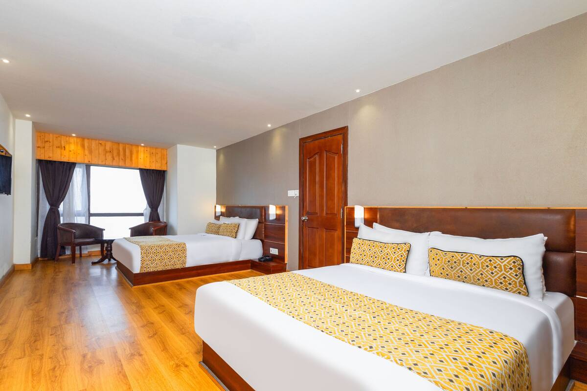 A hotel room with two beds, wooden flooring, a large window with curtains, a wooden door, and a desk with a chair.