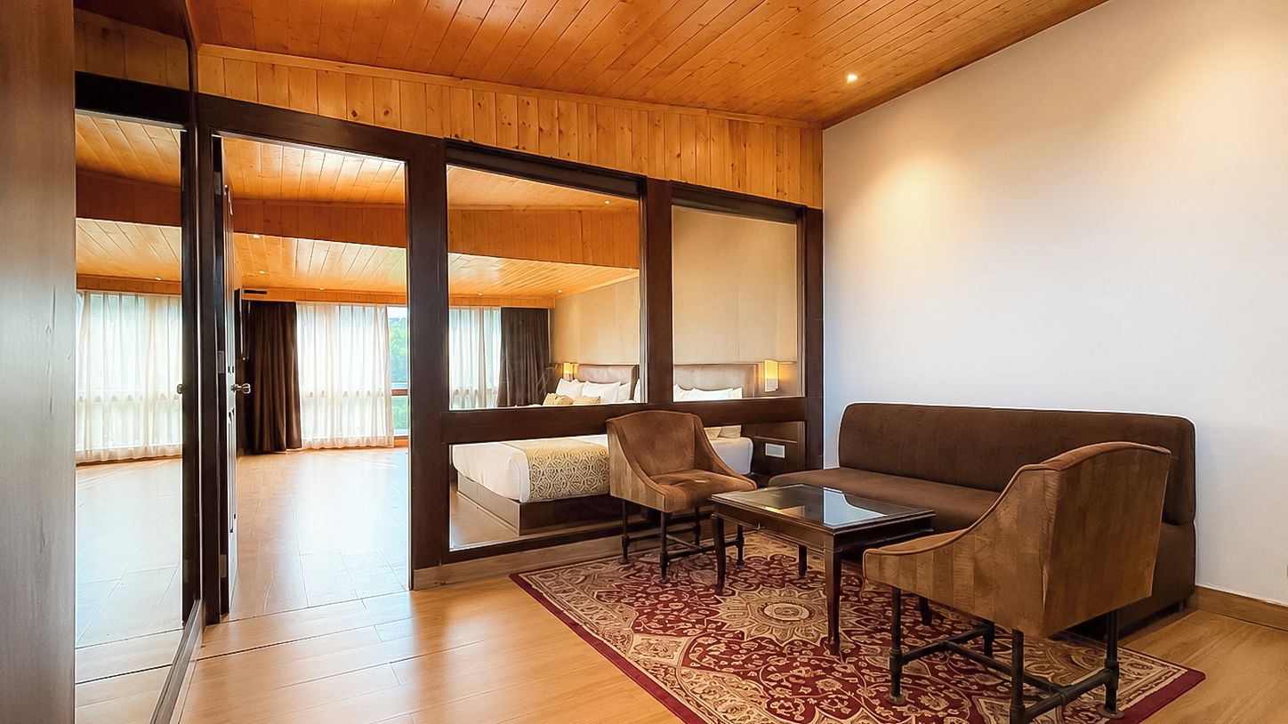 A modern hotel room with a wooden ceiling, a large sliding glass door leading to a balcony, a sofa, two armchairs, and a small table.