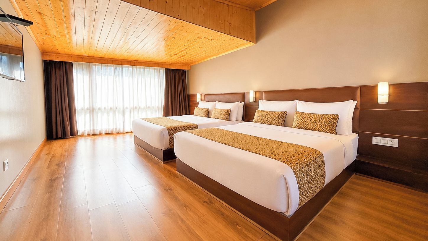 A hotel room with two beds, wooden flooring, a large window with curtains, and a wooden ceiling.