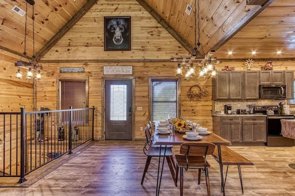 Cozy cabin interior with a high ceiling and exposed beams, featuring a dining area with a wooden table and chandelier, and a kitchen with modern appliances.