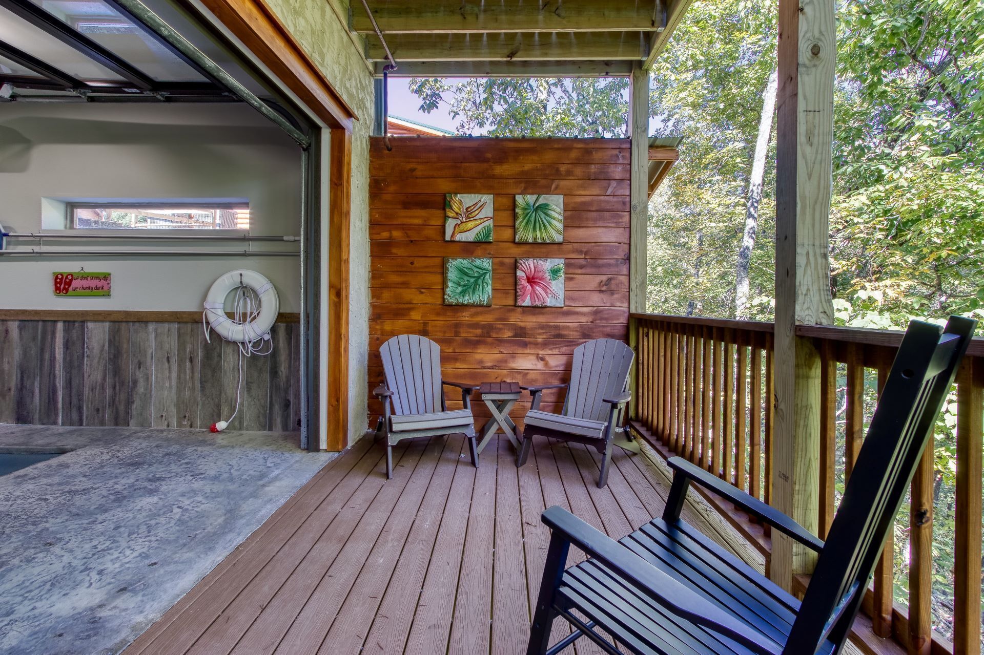 A wooden deck with two Adirondack chairs and a small table, a wooden wall with four paintings, a lifebuoy, and a sign on the wall.
