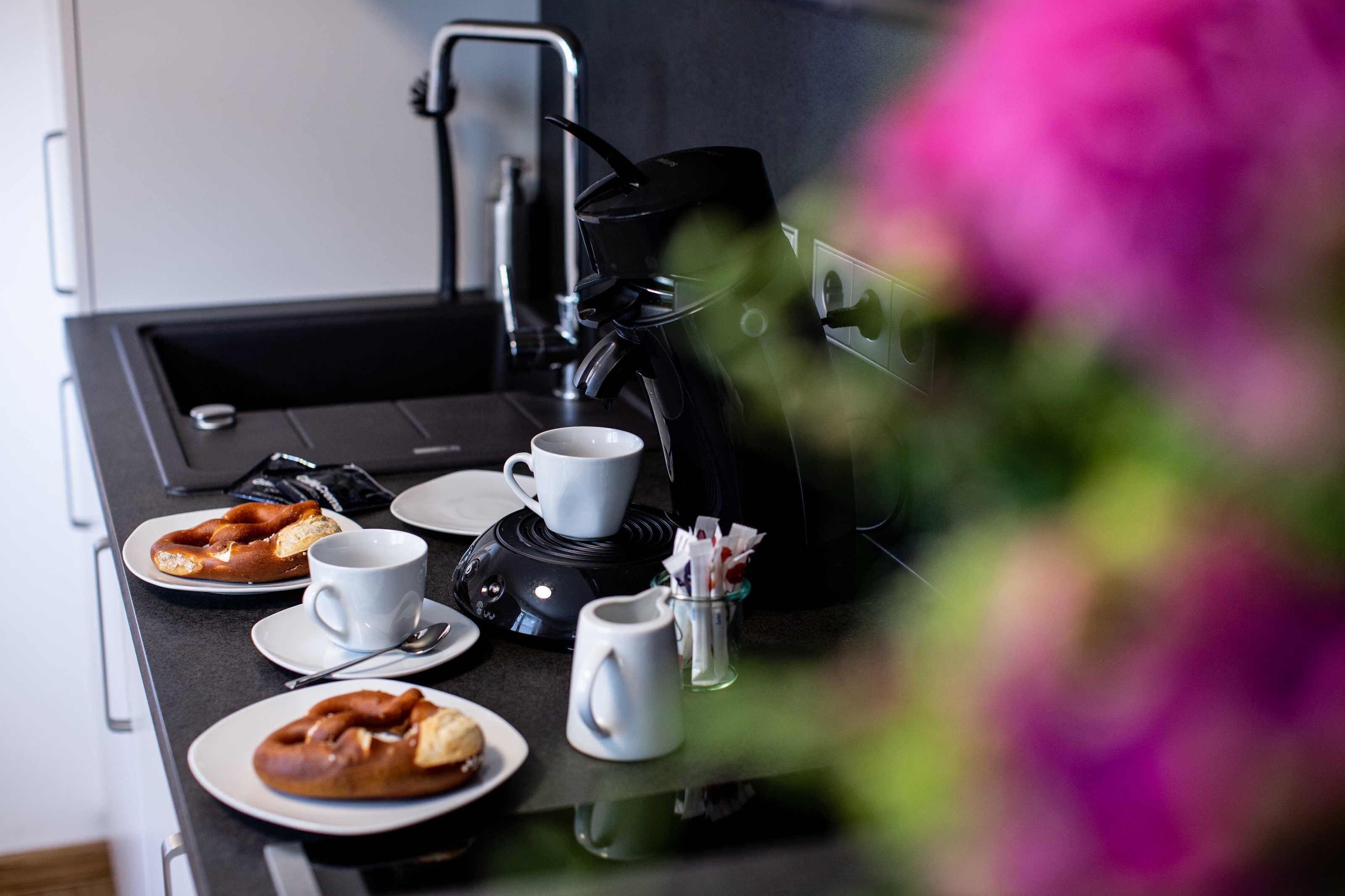 A kitchen counter with a coffee maker, two cups, a plate with a pretzel, and a small pitcher.