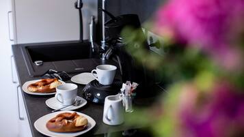 A kitchen counter with a coffee maker, two cups, a plate with a pretzel, and a small pitcher.