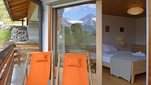 A room with a bed, a window with a mountain view, and two orange chairs.
