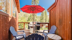 Eine Holzterrasse mit zwei Adirondack-Stühlen, einem Grill und einem roten Sonnenschirm.