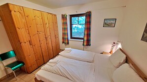 A bedroom with a wooden wardrobe, a bed with white linens, a window with curtains, a radiator, and a framed picture on the wall. - Wohnung 01 EG - Villa Adebar (Heringsdorf)