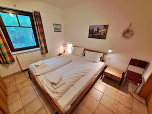 Ein Doppelbett mit weißer Bettwäsche, ein Fenster mit Vorhängen, ein Heizkörper, ein Nachttisch mit Lampe und ein an der Wand hängendes Bild.