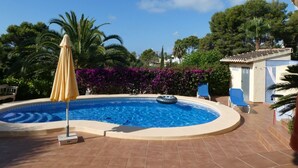 Ein Pool mit runder Form, umgeben von einer gefliesten Liegefläche. Ein gelber Sonnenschirm und eine Liege sind zu sehen.