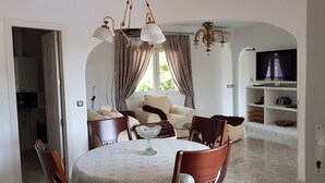 Ein Essbereich mit einem runden Tisch, Stühlen, einer Chandelierleuchtre und einem Fernseher im Hintergrund.
