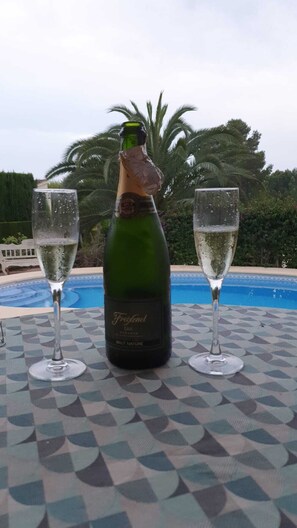 Een fles Brut Nature champagne en twee glazen op een tafel, met een zwembad en palmbomen op de achtergrond.
