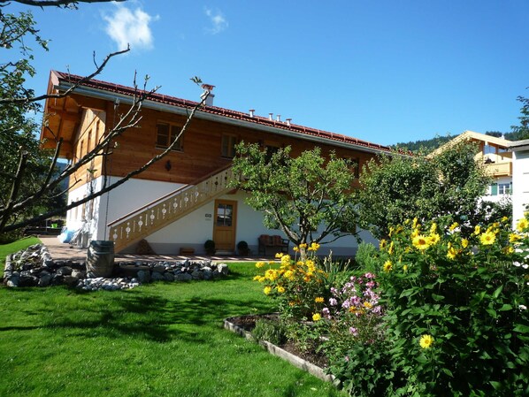 A wooden house with a red-tiled roof, a staircase, and a garden with yellow flowers.