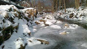 Ein schneebedeckter Fluss mit Steinen, umgeben von kahl stehenden Bäumen und Gebäuden.
