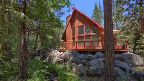 A wooden cabin with large windows, surrounded by rocks and trees.