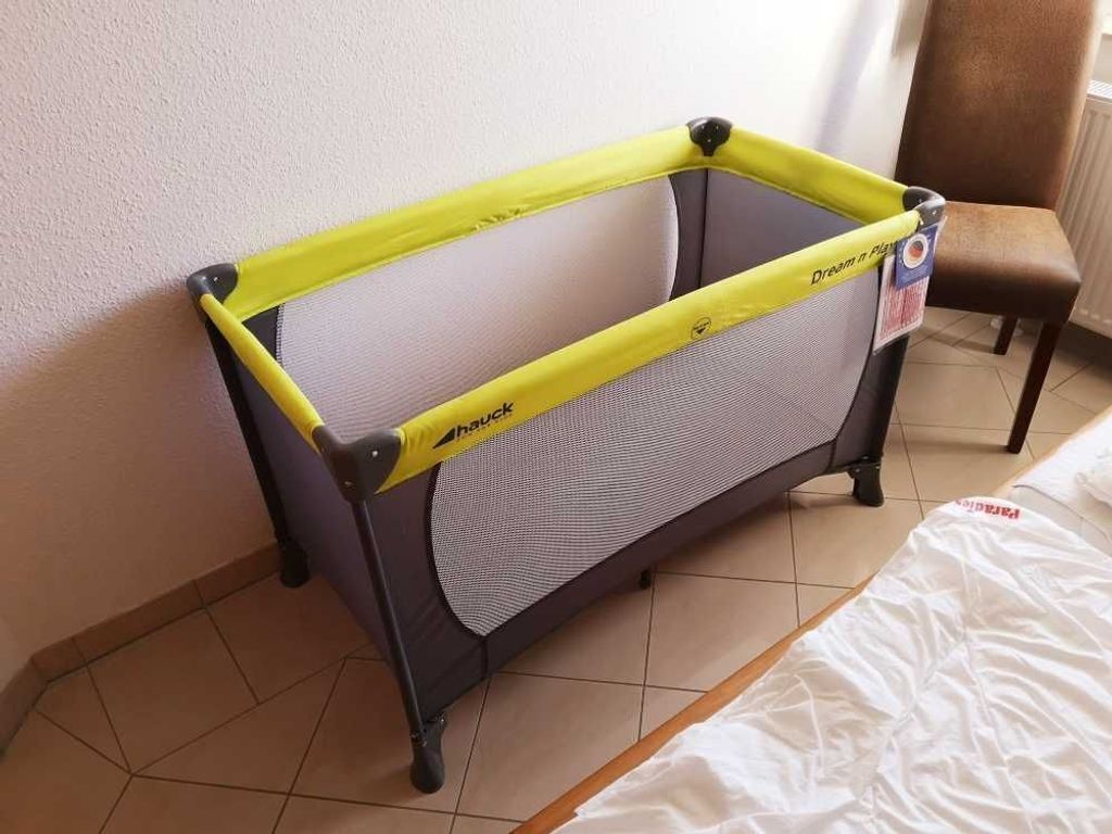 A baby playpen with a yellow frame and a mesh bottom, placed in a corner of a room with a tiled floor and a chair in the background.